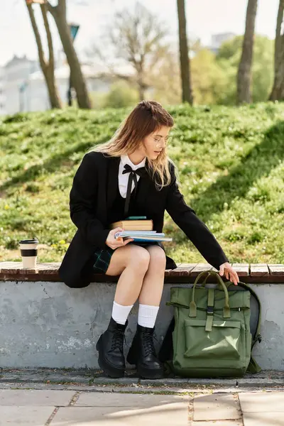 A young adult woman in a chic uniform sits on a wall with her books, sipping coffee. — Stock Photo