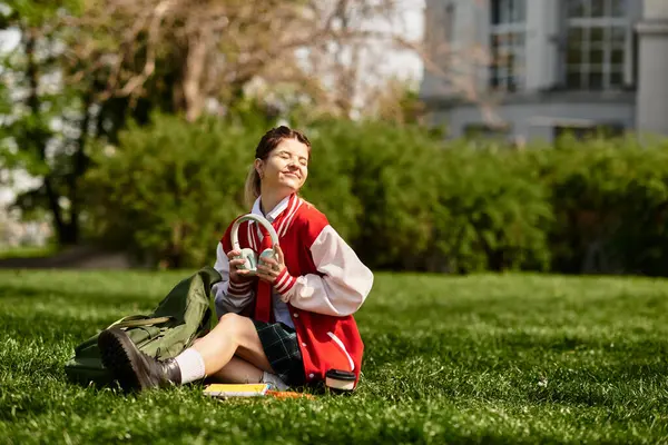 A college girl in a red bomber jacket sits on the grass, smiling with headphones on. — Stock Photo