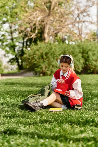 Brillante día en el parque como una mujer joven con una chaqueta de bombardero rojo se sienta en la hierba, estudiando. - foto de stock