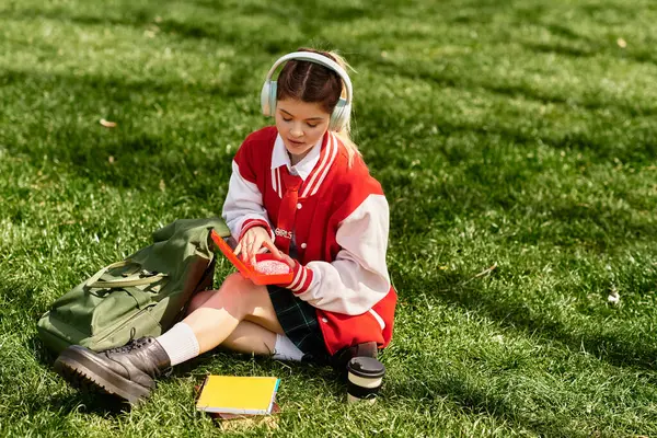 A young college girl sits on the grass, engaged with her phone while dressed in a trendy outfit. — Stock Photo