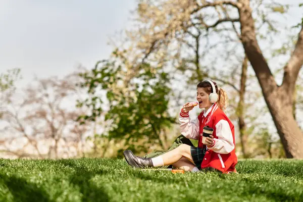 In einem lebhaften Park entspannt sich ein angesagtes College-Mädchen auf dem Rasen und genießt einen Snack und Musik. — Stockfoto
