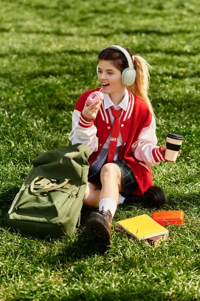College-Mädchen in roter Bomberjacke sitzt im Gras, genießt einen Imbiss und hört Musik. — Stockfoto