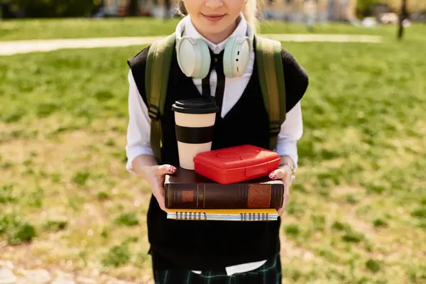 Joven estudiante universitaria se para en el césped, sosteniendo sus libros y café mientras está vestida de moda. - foto de stock