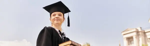 A young woman proudly holds her books while dressed in a graduation gown and cap under a clear sky, banner — Stock Photo