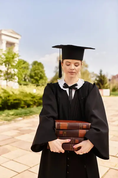 Un graduado en un vestido tiene libros, reflexionando sobre los logros en un campus soleado. - foto de stock