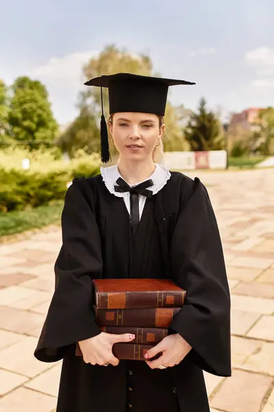 A young graduate in a cap and gown celebrates outside while holding her books. — Stock Photo