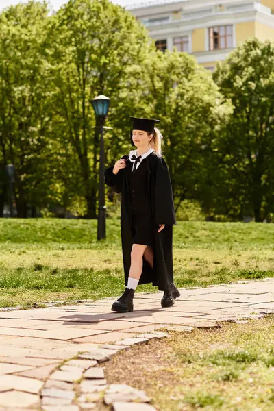 Un giovane adulto in abito da laurea e cappello cammina orgoglioso su un sentiero illuminato dal sole in un ambiente parco. — Foto stock