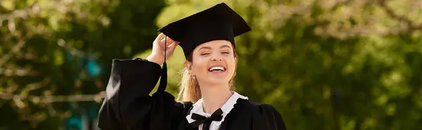 Young college graduate smiles brightly in her gown and cap, banner — Stock Photo