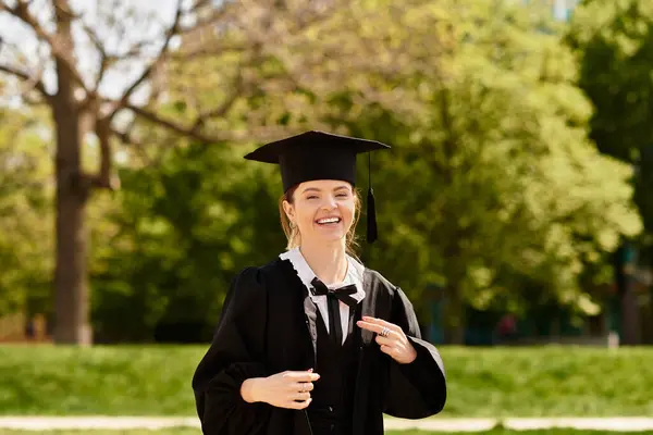 A cheerful college girl in a graduation gown and cap smiles brightly amid lush green scenery. — Stock Photo