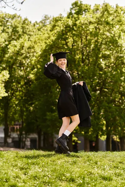 Une jeune fille de l'université dans une robe de graduation et casquette pose joyeusement à l'extérieur entouré de verdure. — Photo de stock