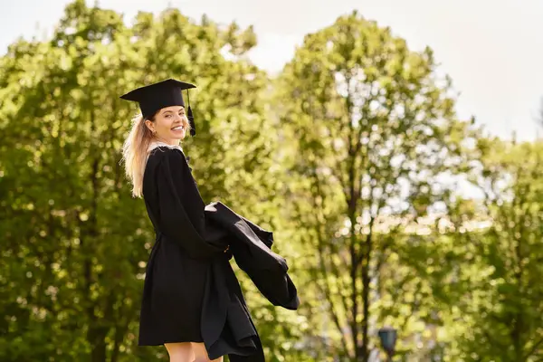 A proud young graduate smiles in her gown and cap, surrounded by trees. — Stock Photo