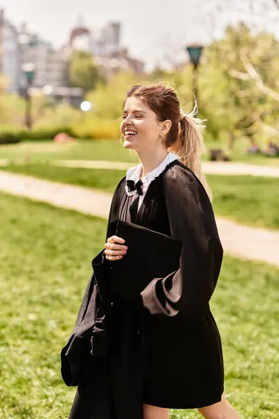 Una giovane donna del college in abito da laurea e cappello sorride con orgoglio mentre cammina in un parco lussureggiante. — Foto stock