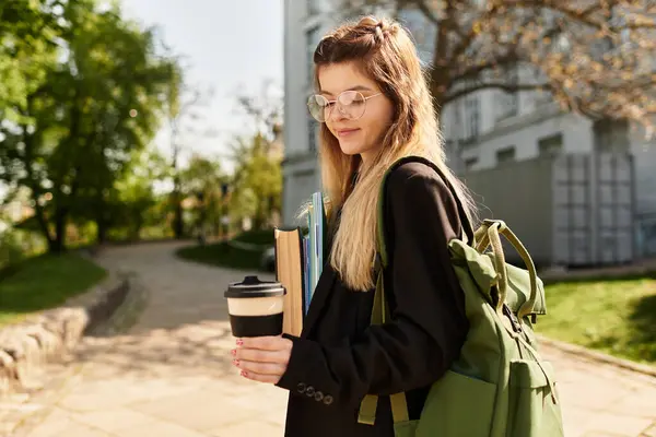 A pretty college girl carries books and coffee while walking through a sunny campus path. — Stock Photo