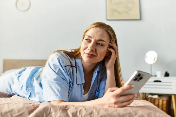 A young woman relaxes on her bed, smiling as she uses her smartphone in a cozy home. — Stock Photo