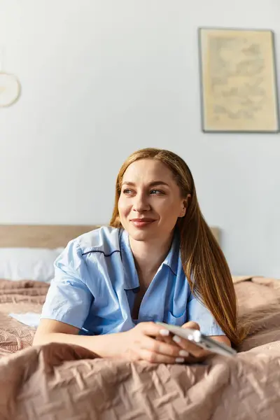 A young woman relaxes on her bed, smiling and holding her phone, enjoying a cozy moment at home. — Stock Photo
