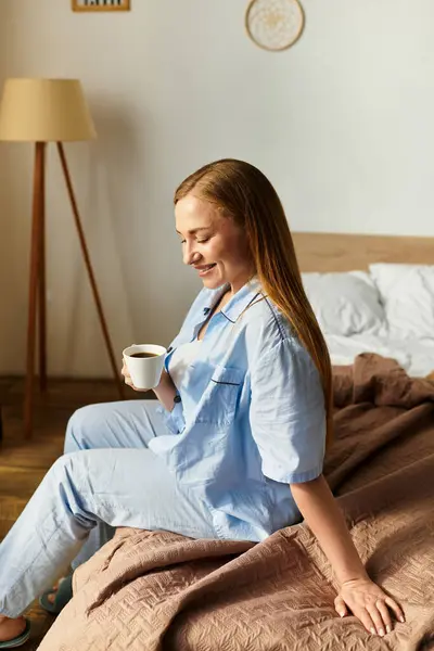 Una giovane donna si siede comodamente sul suo letto, sorseggiando caffè e abbracciando la luce del mattino. — Foto stock