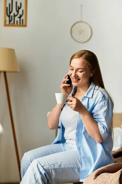 Una bella giovane donna rilassata a casa, chiacchierando gioiosamente al telefono, tenendo in mano una bevanda calda. — Foto stock