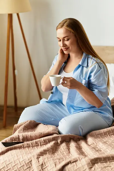 Una giovane donna si siede sul suo letto in pigiama accogliente, assaporando il suo caffè del mattino con un sorriso rilassato. — Foto stock