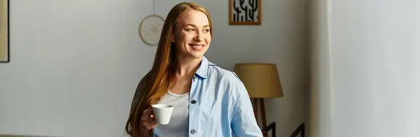 Una mujer joven con el pelo largo se para en casa, felizmente bebiendo su café mientras toma el sol. - foto de stock
