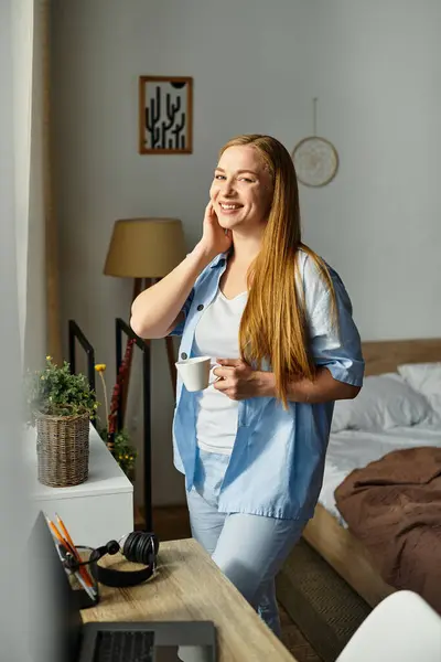 Belle femme se tient près de la fenêtre, souriant alors qu'elle prend un verre chaud dans un cadre confortable à la maison. — Photo de stock