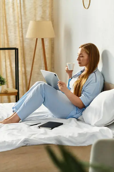 A young woman relaxes on her bed, sipping a hot drink while browsing her tablet in a cozy room. — Stock Photo