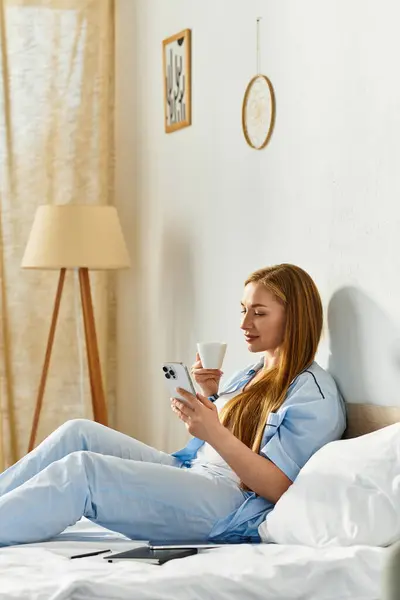 A young woman relaxes at home, sipping a warm drink and checking her phone. — Stock Photo