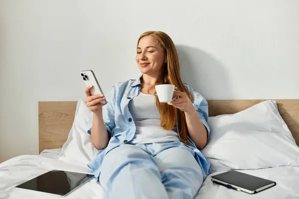 A beautiful young woman relaxes in her cozy bedroom, sipping from a cup and using her phone. — Stock Photo