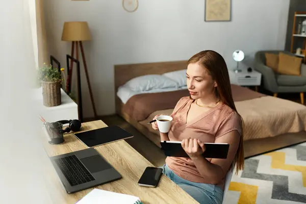 A young woman sits at her desk, sipping a warm drink and focusing on her laptop in a cozy bedroom. — Stock Photo