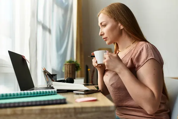 A young woman sips coffee at her desk, concentrating on work in a cozy home. — Stock Photo
