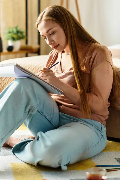 A young woman sits on the floor, sketching in her notebook, lost in creativity at home. — Stock Photo