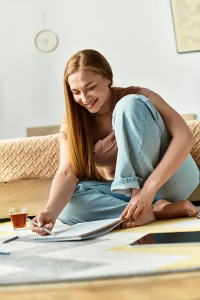 A beautiful young woman sits comfortably on the floor, focused on her drawing with tea beside her. — Stock Photo