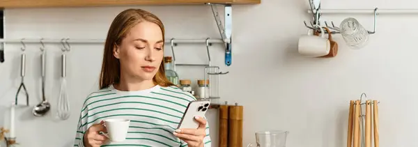 A young woman relaxes at home in her kitchen, holding a cup of coffee and looking at her phone. — Stock Photo