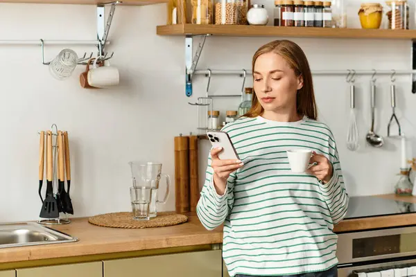 Young woman relaxes in her cozy kitchen, sipping coffee while looking at her phone. — Stock Photo