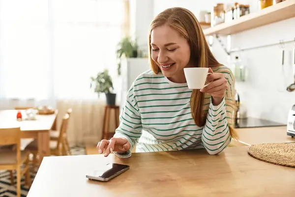 A young woman smiles while sipping a warm beverage and using her smartphone in a cozy kitchen. — Stock Photo