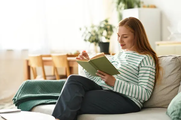 Молодая женщина комфортно отдыхает на диване, глубоко занятая в своей книге в уютной домашней обстановке. — стоковое фото