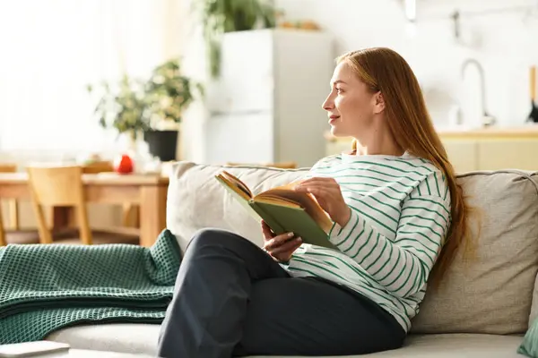 Young woman seated comfortably on a couch, deeply engaged in reading a book in her serene home. — Stock Photo