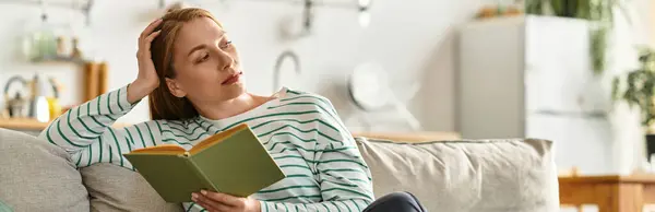 A beautiful young woman sits comfortably on a sofa, immersed in a book while relaxing at home. — Stock Photo