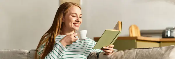 A young woman smiles while reading a book and sipping coffee in a warm and inviting home atmosphere. — Stock Photo