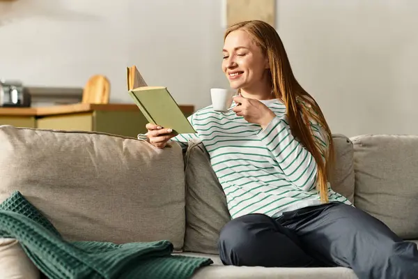 Una giovane donna si siede comodamente sul suo divano, sorseggiando tè mentre legge un libro nella sua accogliente casa. — Foto stock