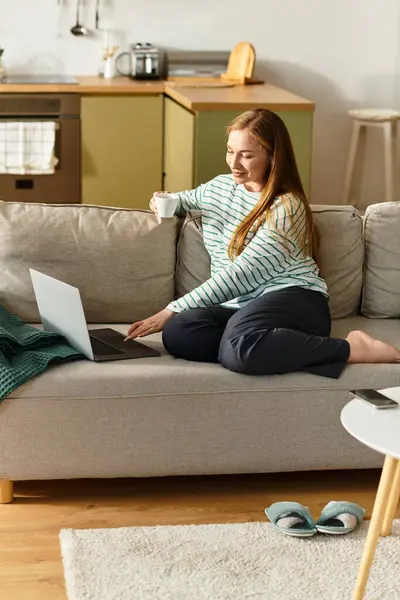 Rilassandosi a casa, una giovane donna si siede su un divano, usando il suo computer portatile e gustando una calda tazza di tè.. — Foto stock