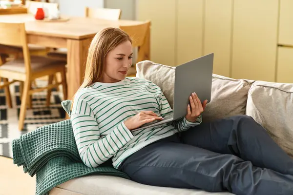 A beautiful woman relaxes on the couch, deeply engaged with her laptop in a warm atmosphere. — стоковое фото