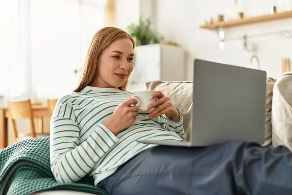 Relaxed and beautiful woman sits comfortably at home, sipping a warm drink and engaged online. — Stock Photo