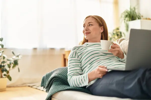 Una giovane donna si rilassa a casa sorseggiando un drink caldo mentre usa il suo portatile. — Foto stock