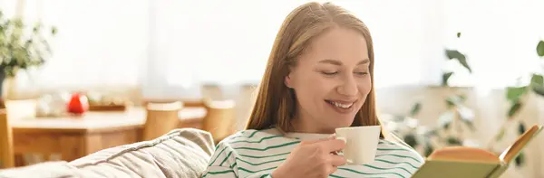 A young woman smiles as she sips her warm drink while reading a book in her cozy living space. — Stock Photo