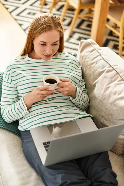 Beautiful woman relaxes at home with coffee, immersed in her laptop on a comfy couch. — Stock Photo
