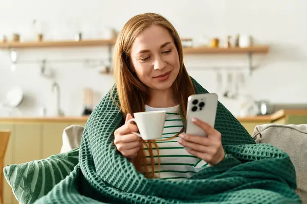 Una giovane donna avvolta in una coperta calda si gode una tazza di caffè mentre usa il suo smartphone a casa. — Foto stock