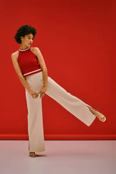 A striking young woman poses elegantly with one leg raised in trendy attire against a red backdrop. — Stock Photo