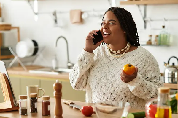 A young African American woman happily cooks a salad, engaged in a phone conversation at home. — Stock Photo