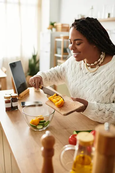 Uma jovem mulher negra sorri enquanto ela chops legumes frescos para uma salada colorida em sua cozinha acolhedora. — Fotografia de Stock