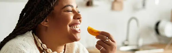 Uma jovem afro-americana comendo tomate com entusiasmo em sua cozinha aconchegante, banner — Fotografia de Stock